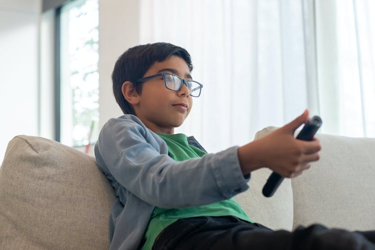 A young boy sits on a couch with a TV remote.