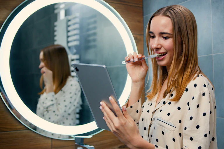 A young woman brushing her teeth while looking at a tablet screen.