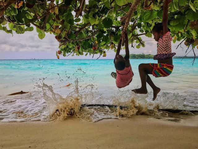 Children hanging from a tree overhanging a tropical beach as the surf breaks.