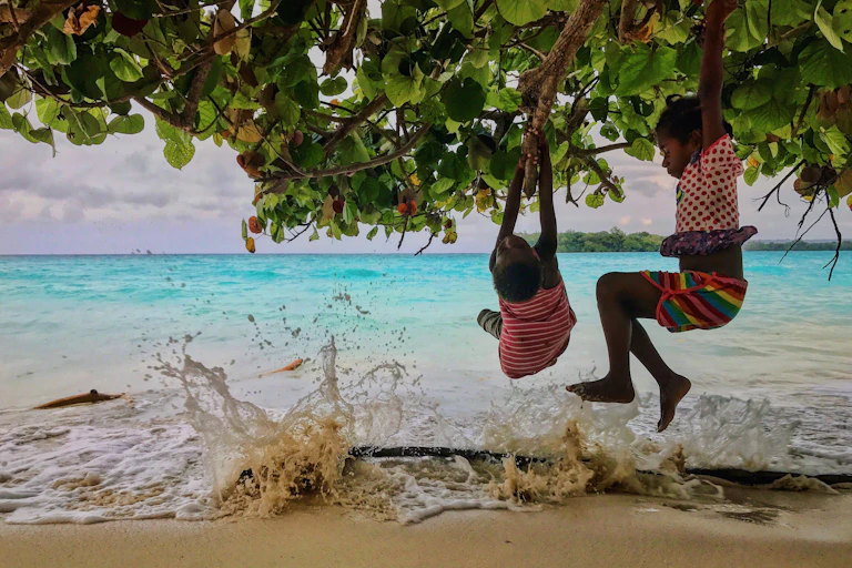 Children hanging from a tree overhanging a tropical beach as the surf breaks.