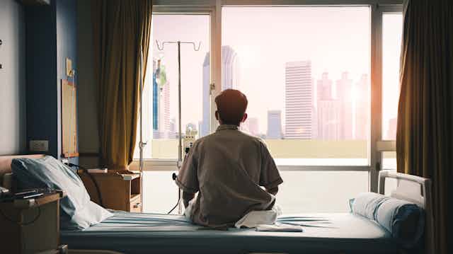 A male patient sits alone in a hospital room looking out the window.