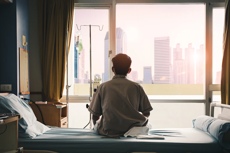 A male patient sits alone in a hospital room looking out the window.