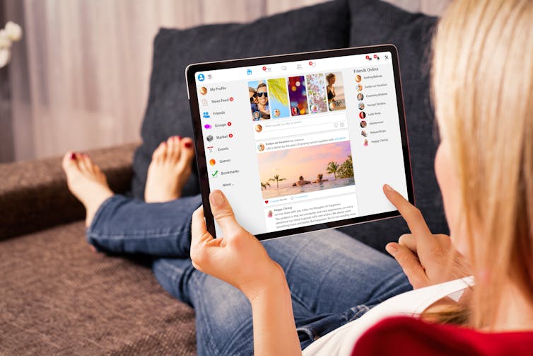 a woman sits on a couch and scrolls through Facebook on a tablet