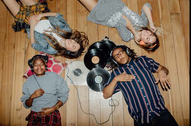 Four young people lying on tan hardwood floor listening to music with over-the-ear headphones, and vinyl records strewn on the floor. 