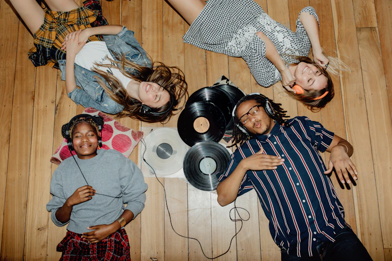 Four young people lying on tan hardwood floor listening to music with over-the-ear headphones, and vinyl records strewn on the floor.