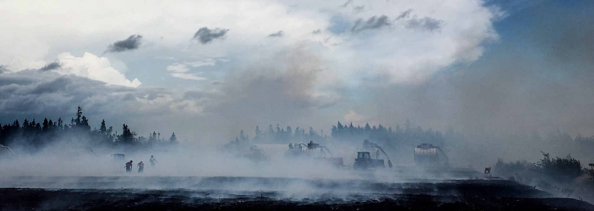 Smoke rises above a field with people and vehicles.