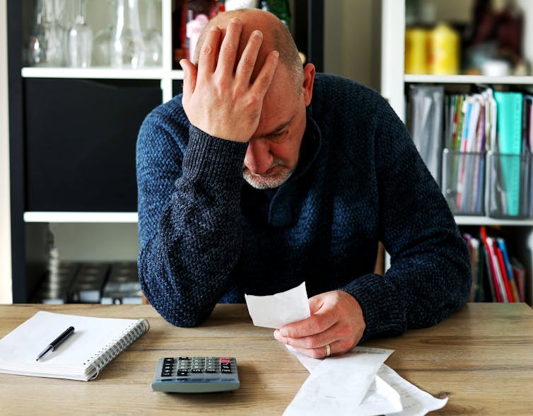 An older man sits at a table with his head in his head as he reads a receipt