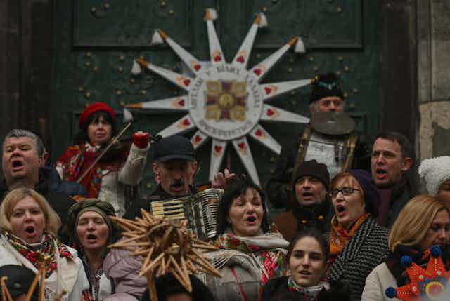 About a dozen people in winter clothing sing enthusiastically outside a large green door with a white star decoration.