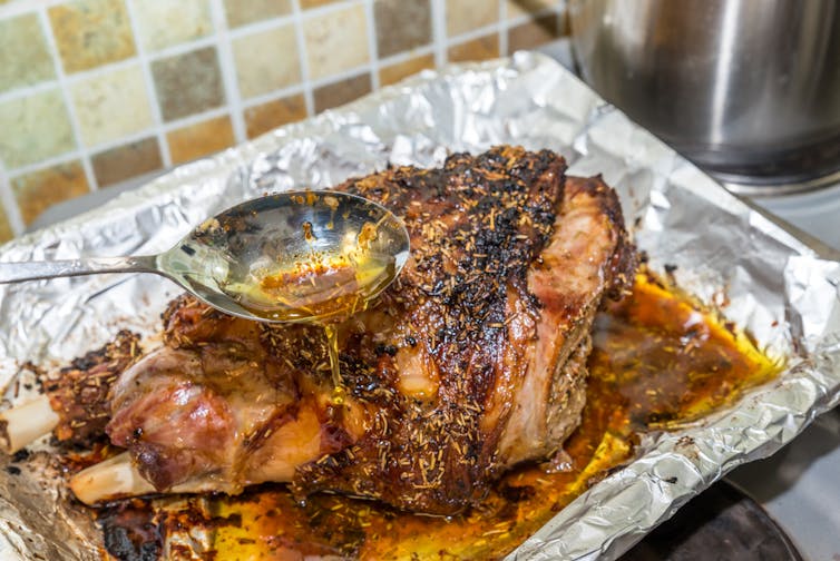 Meat juices drip off a spoon into a tray of roasted meats.