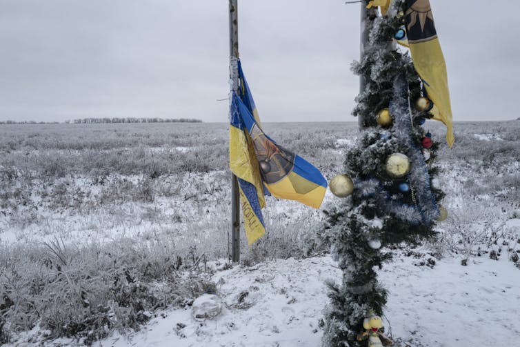 A blue and yellow flag flutters on a short flagpole on a snow-covered field, next to a thin tree decorated with a few ornaments.