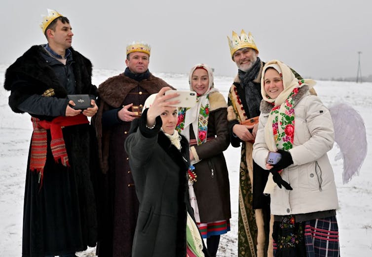 Six people in heavy coats and festive crowns stand outside, taking a selfie on a smart phone, in a snow-covered field.