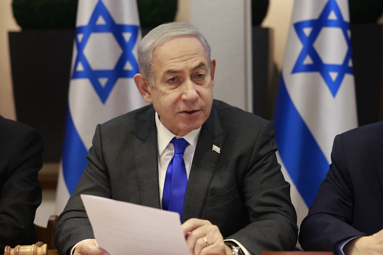 A man with grey hair in a suit talks in front of two Israeli flags