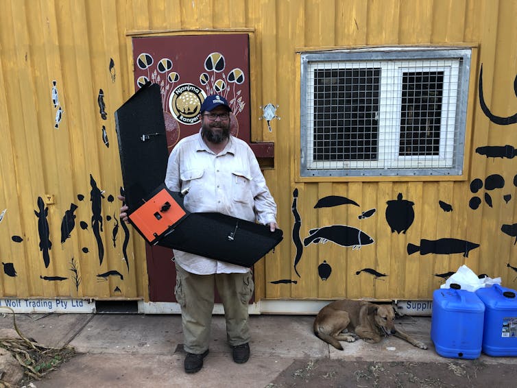 A bearded smiling man holding a large black and red device with wings