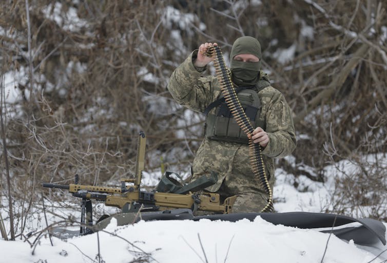 A man in military uniform and wearing a balaclava loads bullets into an automatic rifle.