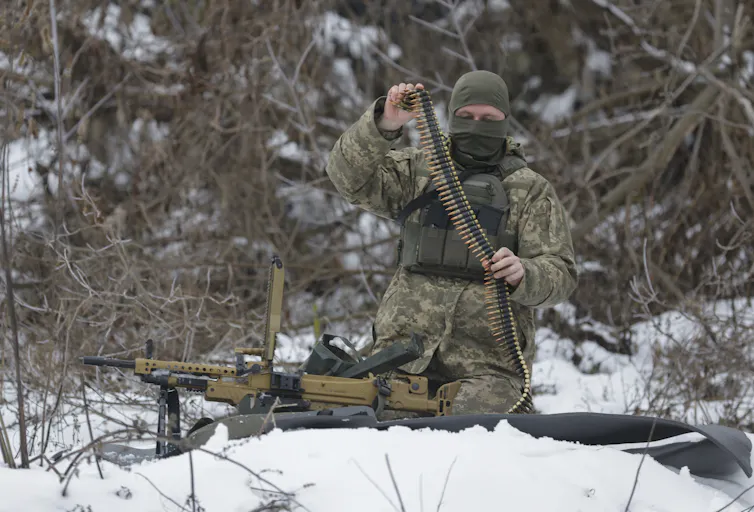 A man in military uniform and wearing a balaclava loads bullets into an automatic rifle.