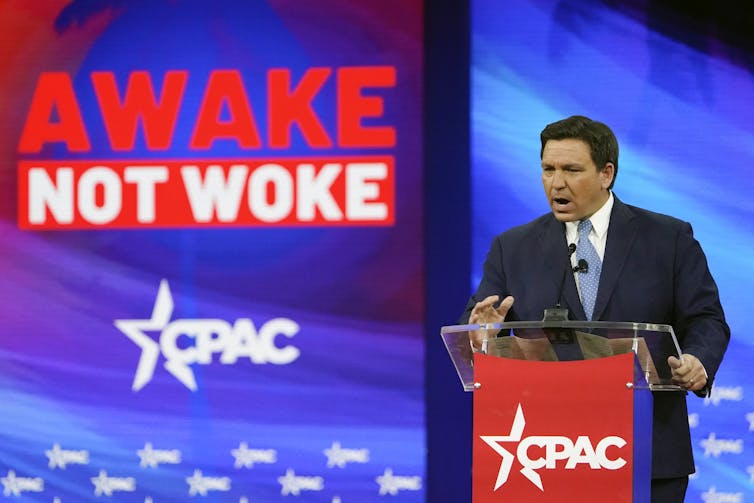 A dark-haired man speaks into a microphone in front of a sign that reads Awake Not Woke.