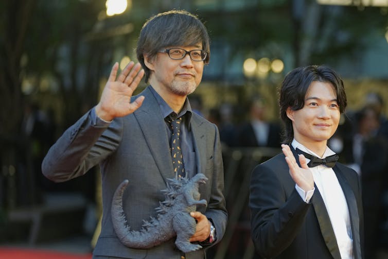 Two men in suits, one holding a large plastic lizard, are waving.