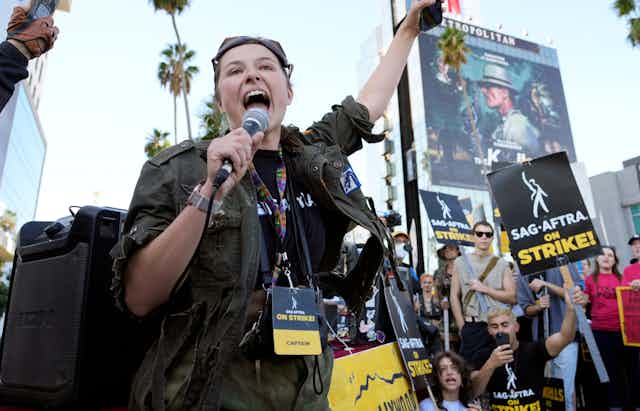 A woman leads chanting on a picketline surrounded by people waving signs saying 'SAG AFTRA on strike!'