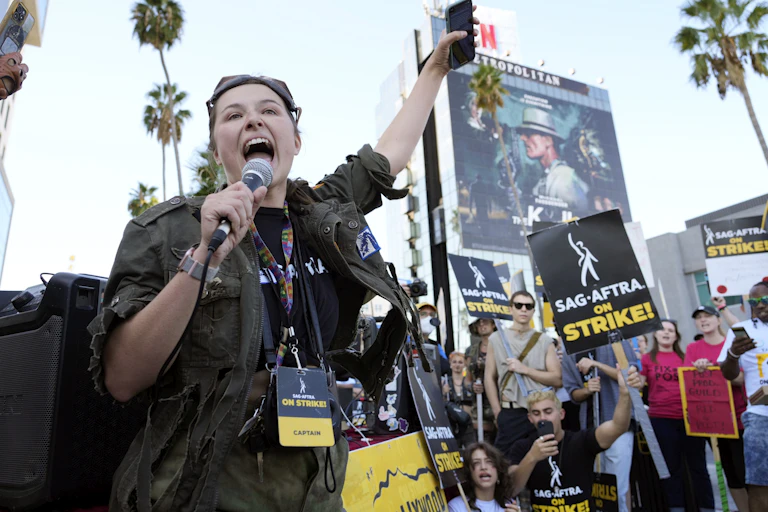 A woman leads chanting on a picketline surrounded by people waving signs saying 'SAG AFTRA on strike!'
