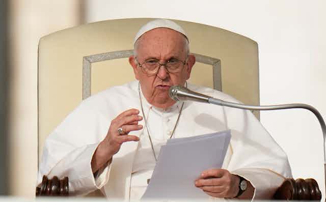 Pope Francis in white priestly garments, holding a sheaf of papers, as he speaks at the lectern.