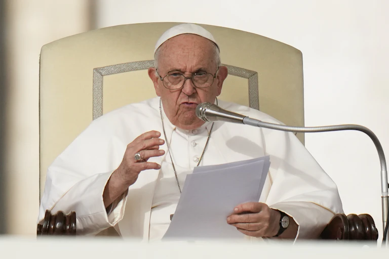 Pope Francis in white priestly garments, holding a sheaf of papers, as he speaks at the lectern.