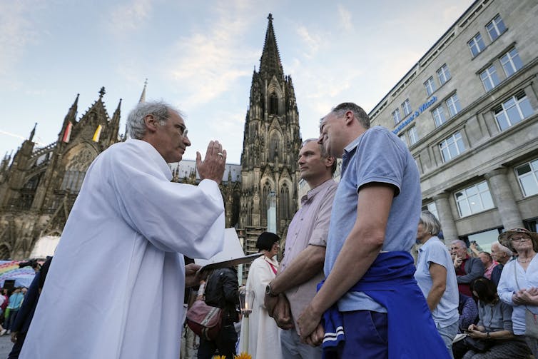 Two people stand in front of a cathedral while another man in white priestly garments blesses them.