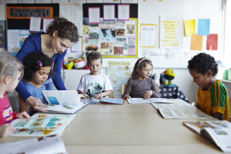 Five students sit at a table doing work under as a teacher checks their work.