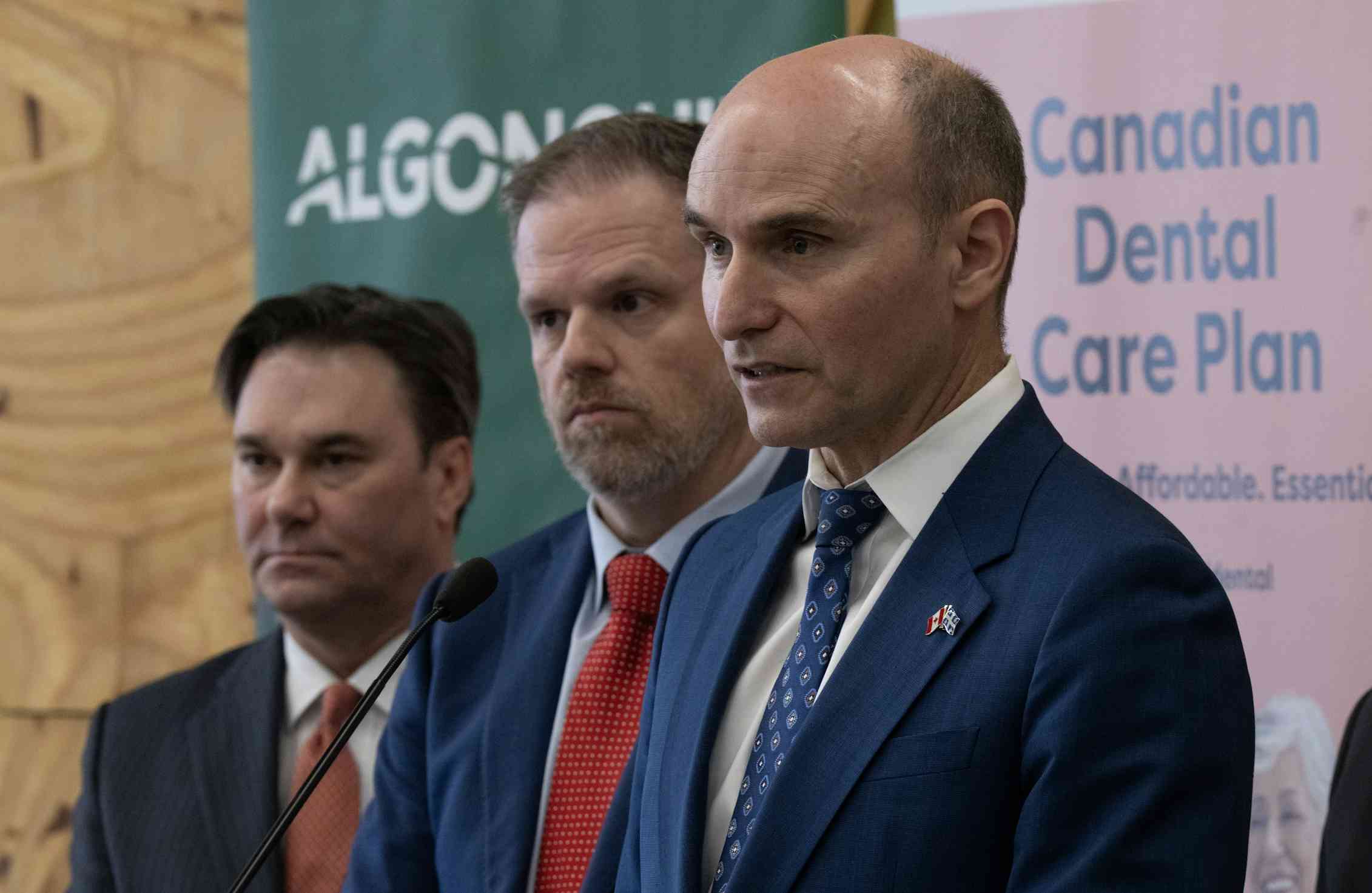 Three men in dark suits in front of a sign reading 'Canadian Dental Care Plan'