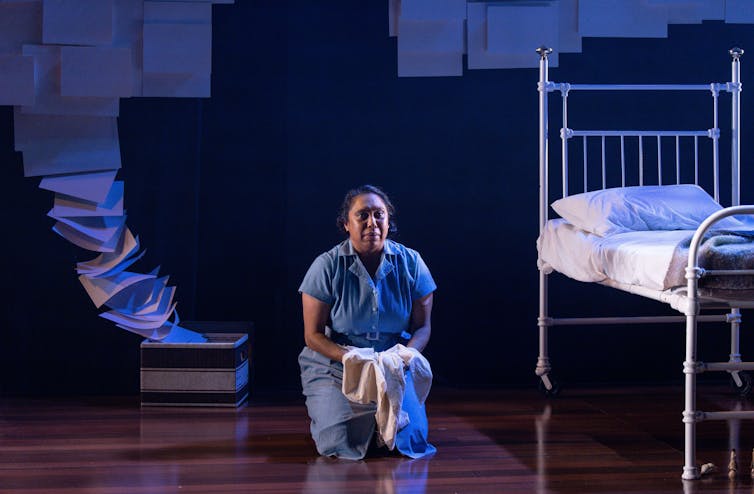 A woman dressed as a maid, near a white metal bed.