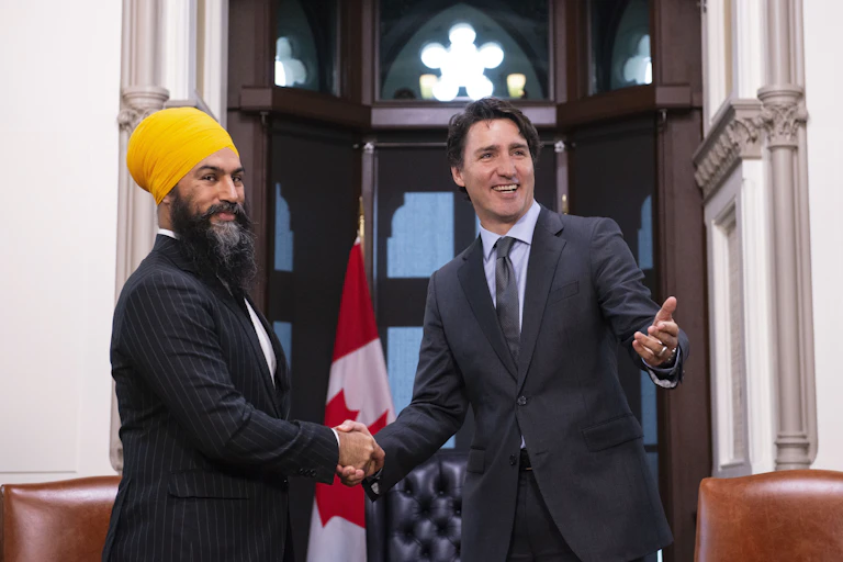 Two men, one in a yellow turban, smile as they shake hands. A Canadian flag is behind them.