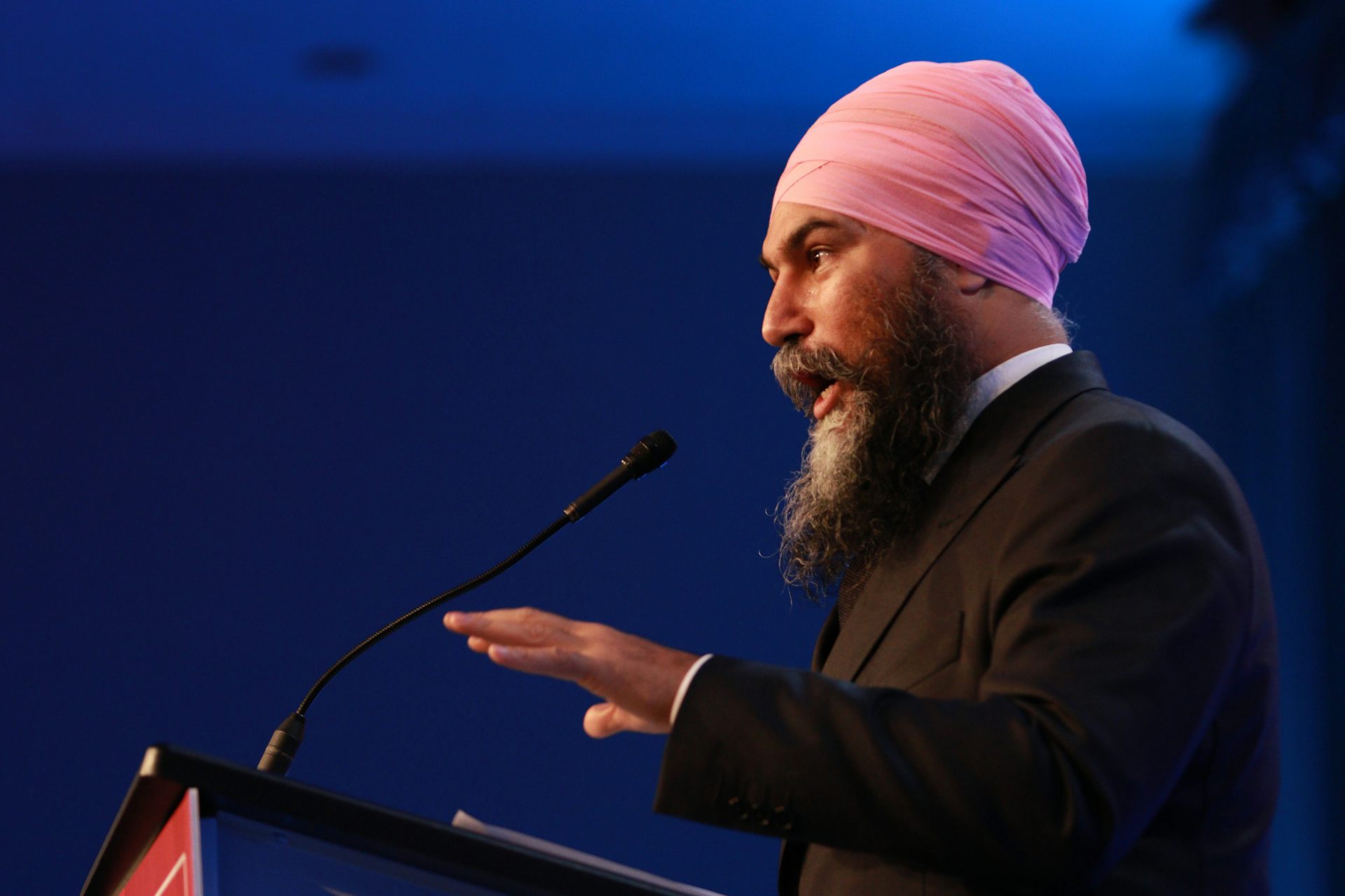 A bearded man in a pink turban speaks from behind a podium.