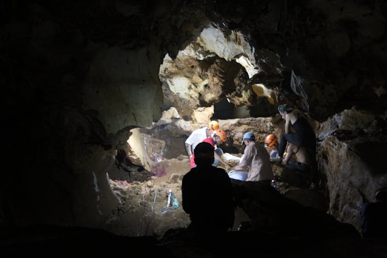 Uma caverna escura com paredes claras, com várias pessoas com capacetes olhando através do sedimento