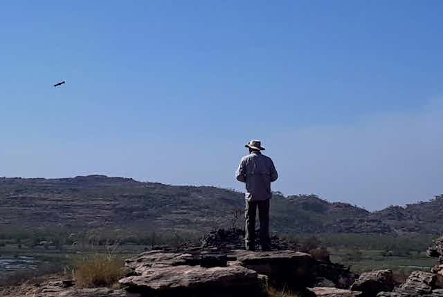 A person with a wide brimmed hat standing on a rocky landscape operating a drone in a blue sky