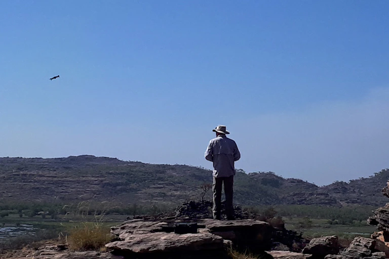 A person with a wide brimmed hat standing on a rocky landscape operating a drone in a blue sky
