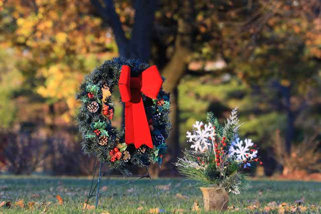 Holiday wreath with a large red bow next to a holiday floral arrangement at a gravesite. 