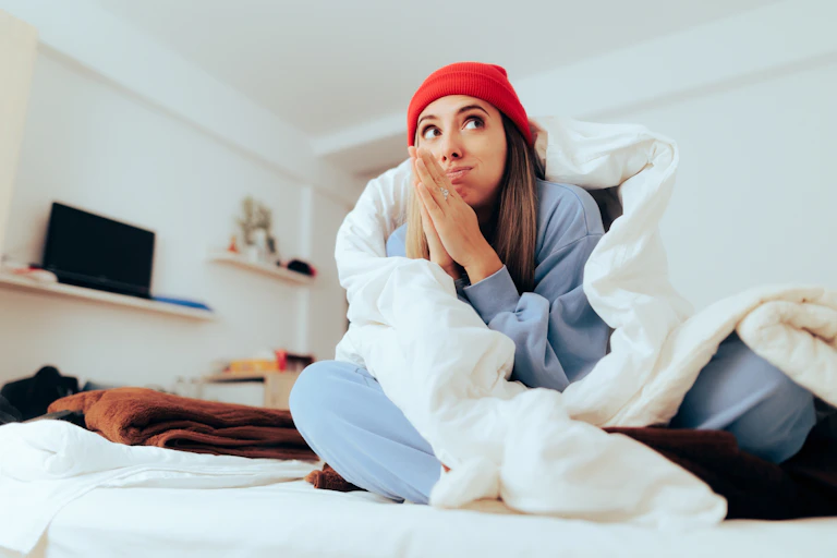 Woman in red hat huddles under a blanket and rubs her hands together