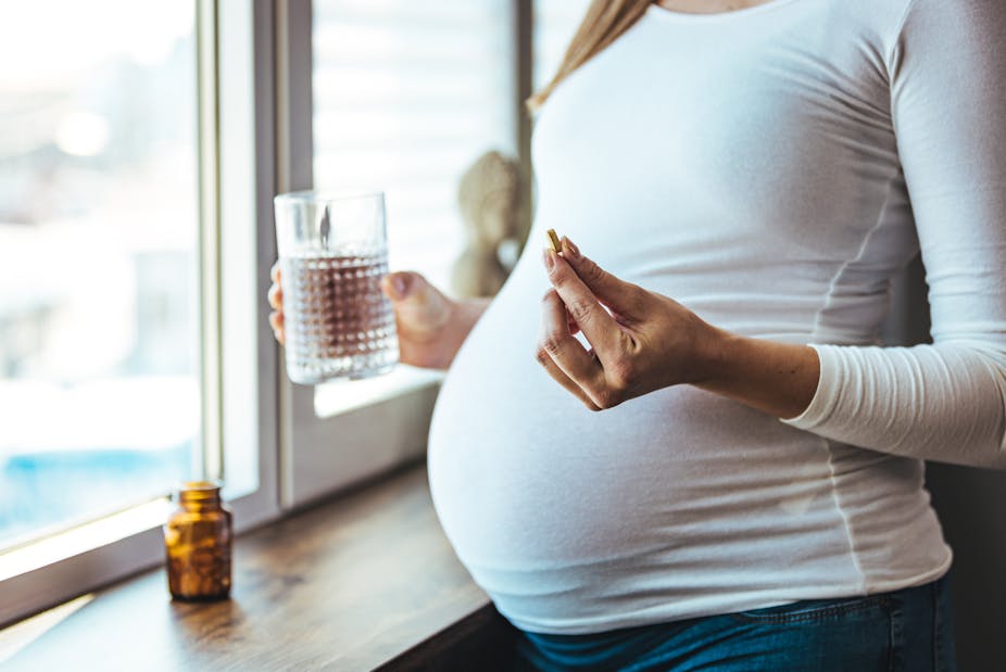 A pregnant woman holds a glass of water in one hand and a vitamin supplement in the other.