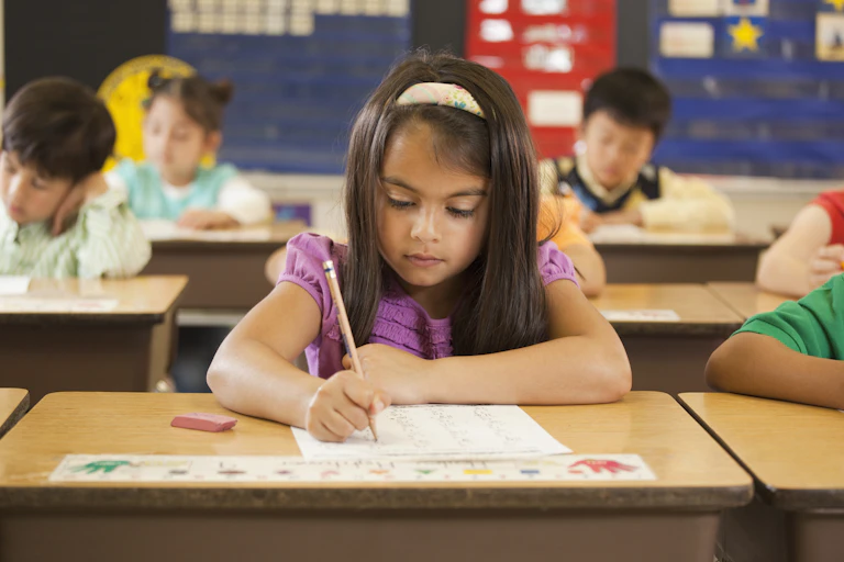 Young girl sits at school desk surrounded by classmates taking a test