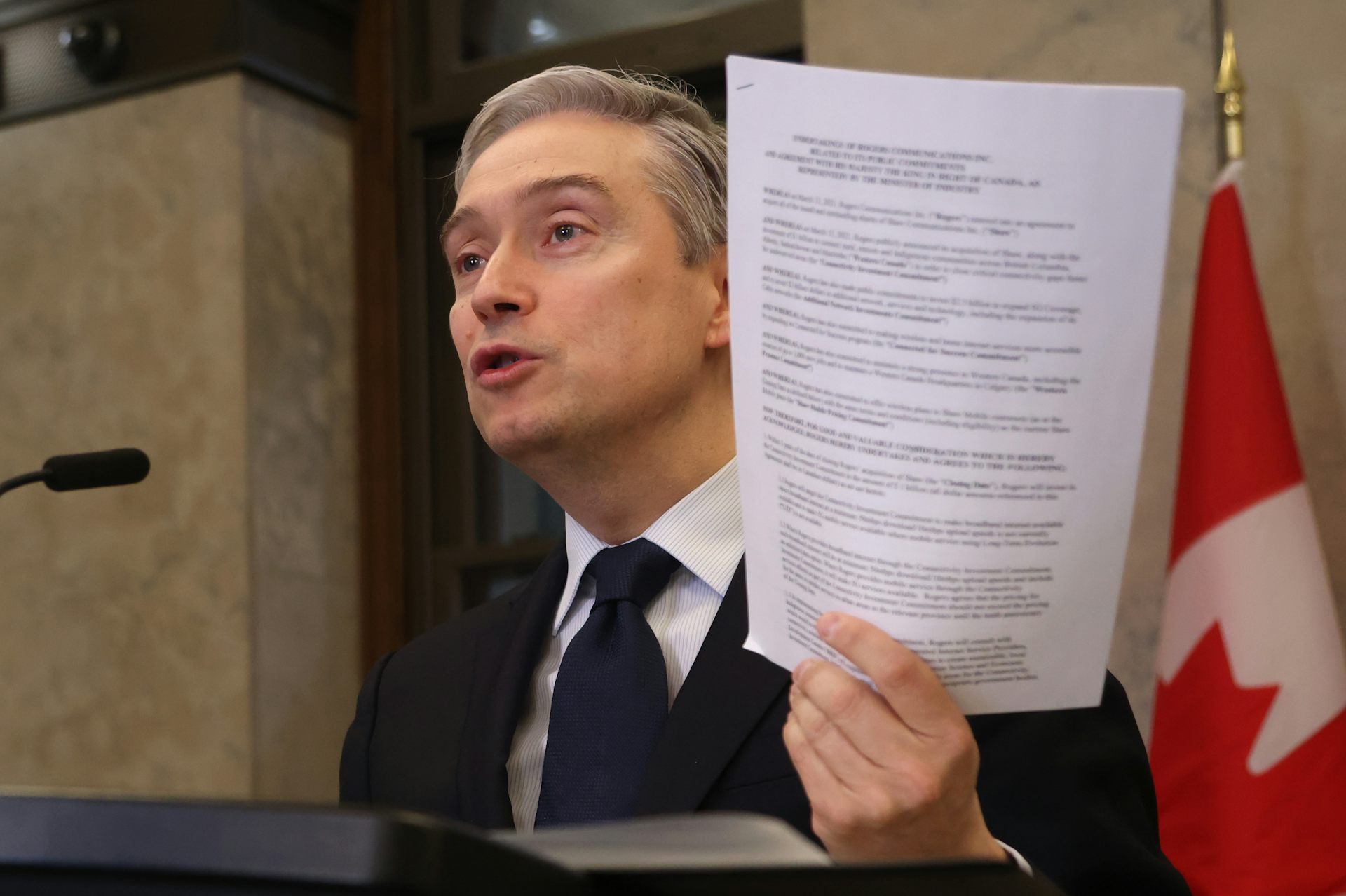 A middle-aged man speaking into a microphone on a podium while holding up a stack of papers