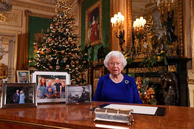 An old lady, the Queen, dressed in blue, sitting at a desk in front of large Christmas tree.