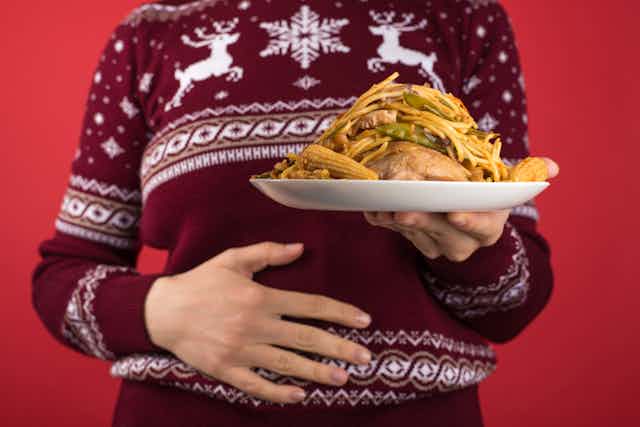 Close-up of person in holiday sweater holding a plate full of food with one hand on their stomach