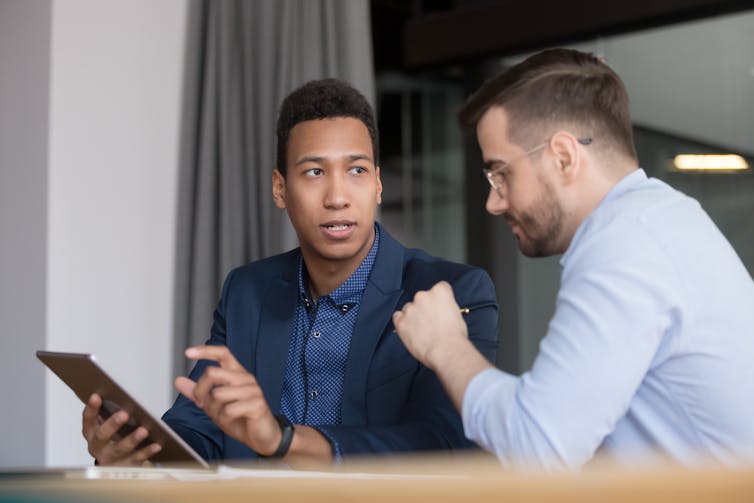 Two men in business attire have a conversation while seated at a table. One is holding a tablet.