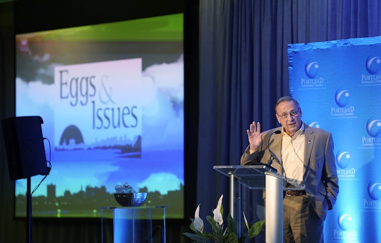 A big sign projected on a wall that says 'Eggs & Issues' with a man to the right at a lectern, talking.