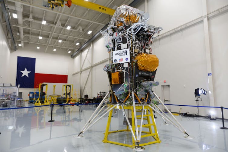 A white room with a Texas flag in the background and a bundle of machinery, sitting on a yellow stand, in the foreground.