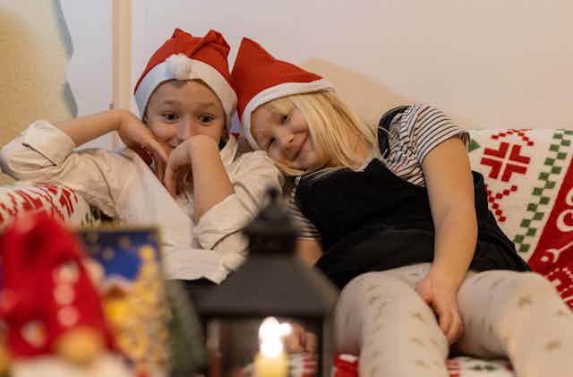 Two kids sit on a couch with Santa hats looking very happy. 