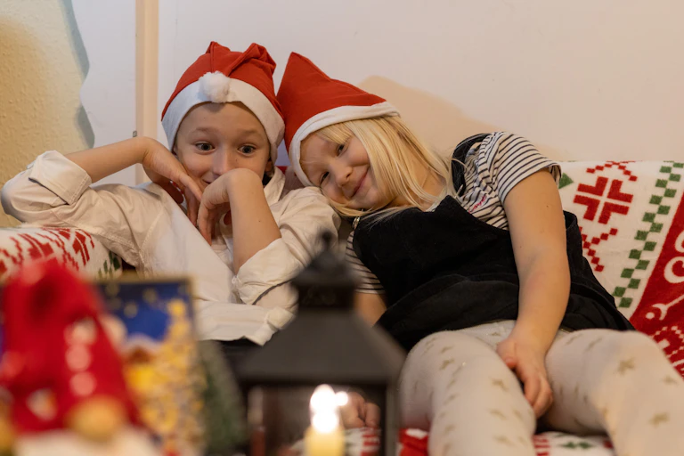 Two kids sit on a couch with Santa hats looking very happy.