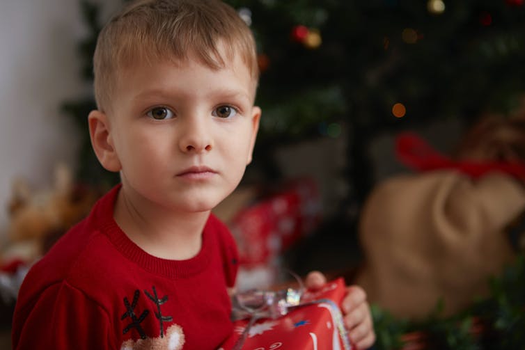 A young child holds a gift in front of the tree, looking very serious.