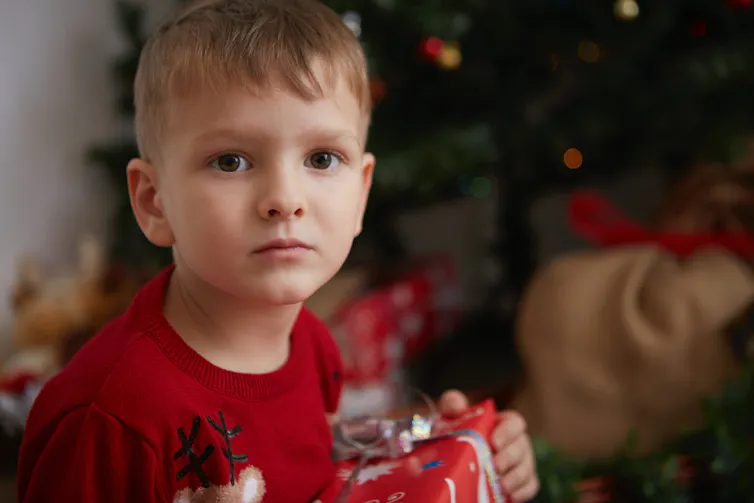 A young child holds a gift in front of the tree, looking very serious.