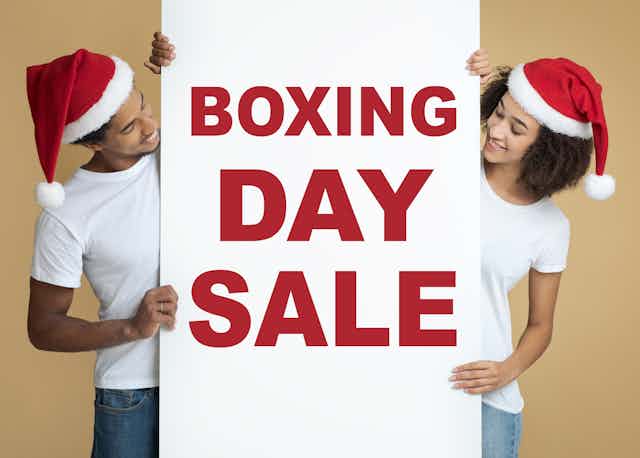 Man and woman wearing Santa hats holding up a "Boxing Day Sale" sign