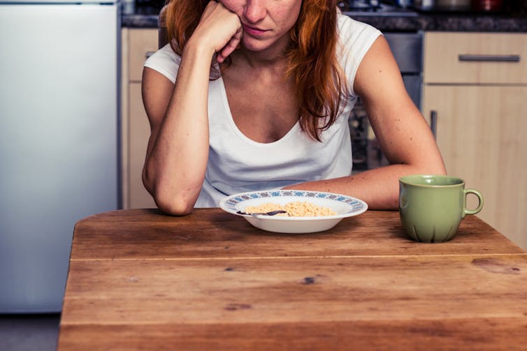 زن خسته که به کاسه صبحانه، غلات و فنجان روی میز آشپزخانه خیره شده است